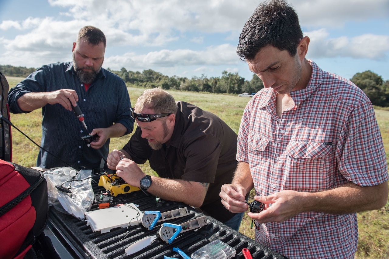 Three men use hand tools on small electronic devices in a field.