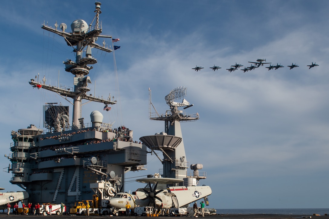 Eleven aircraft fly in formation as dozens of sailors watch from the bridge of an aircraft carrier.