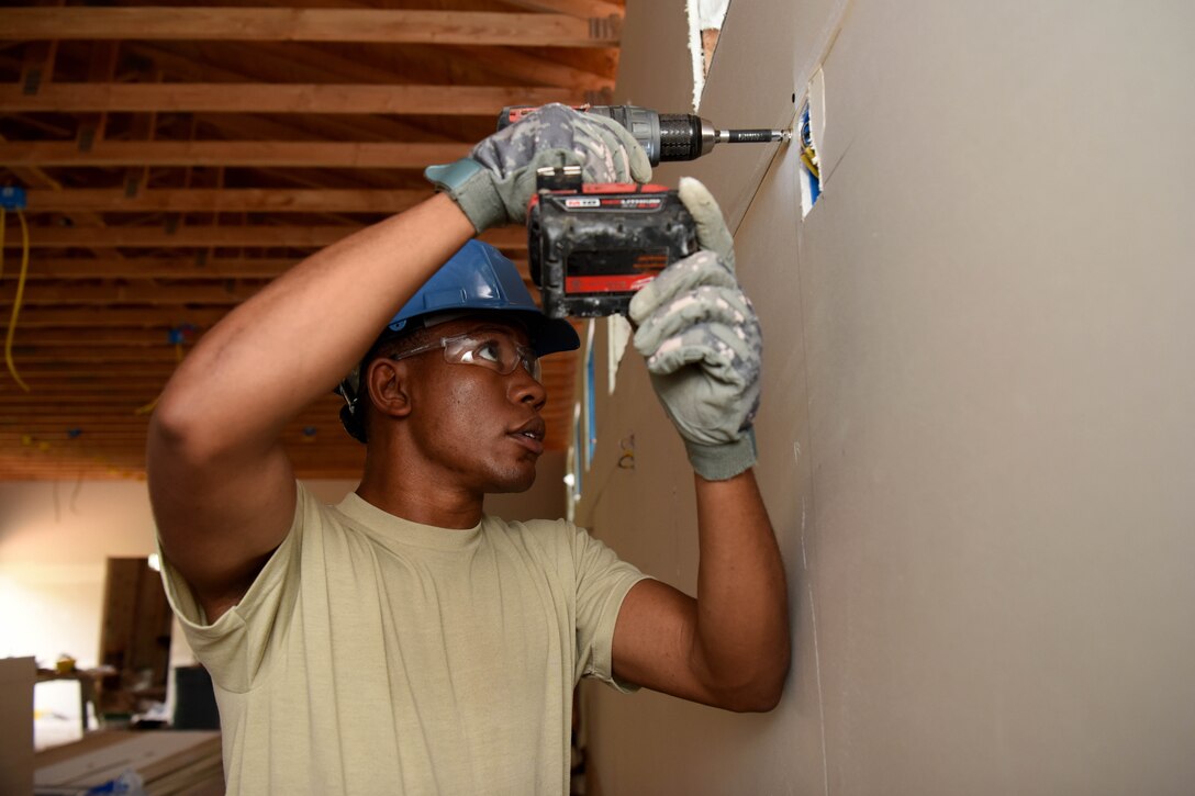 An airman wearing a hard hat, goggles and gloves drills a hole into sheetrock.