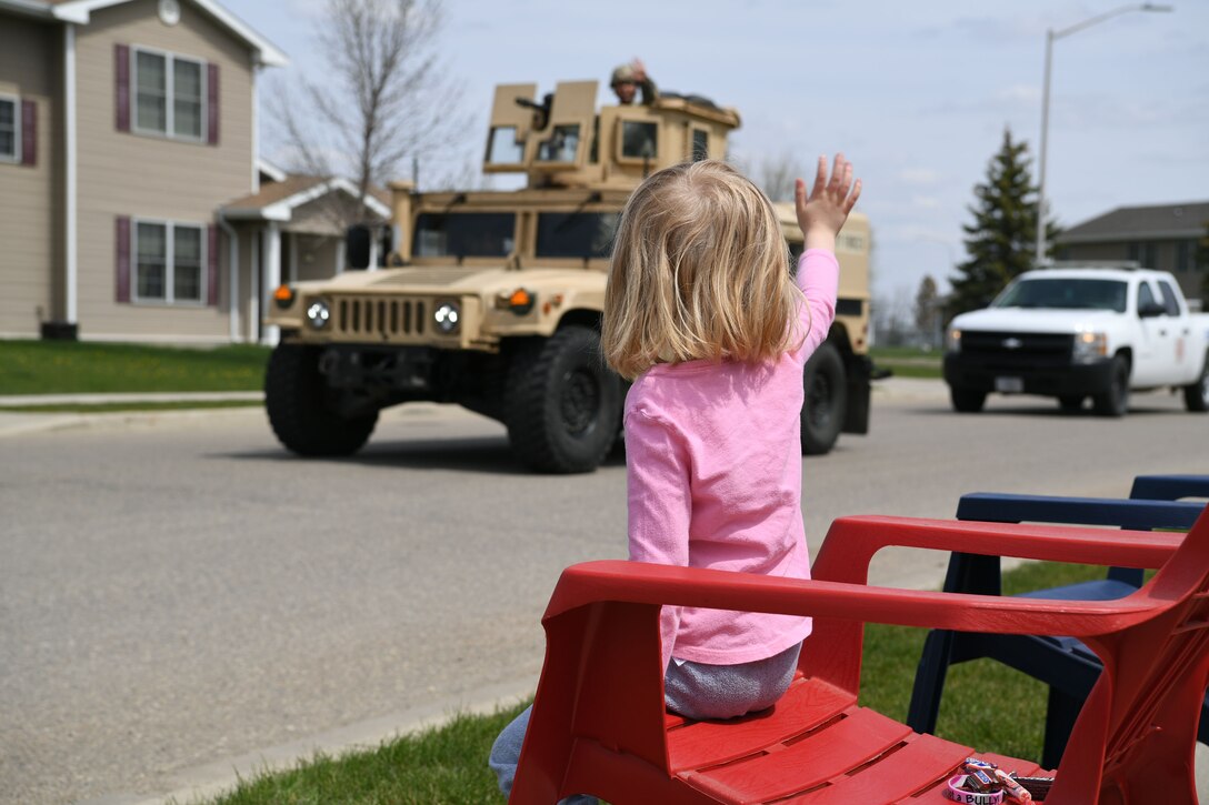 A child in a red lawn chair waves as an airman in a Humvee passes, followed by a pickup truck.