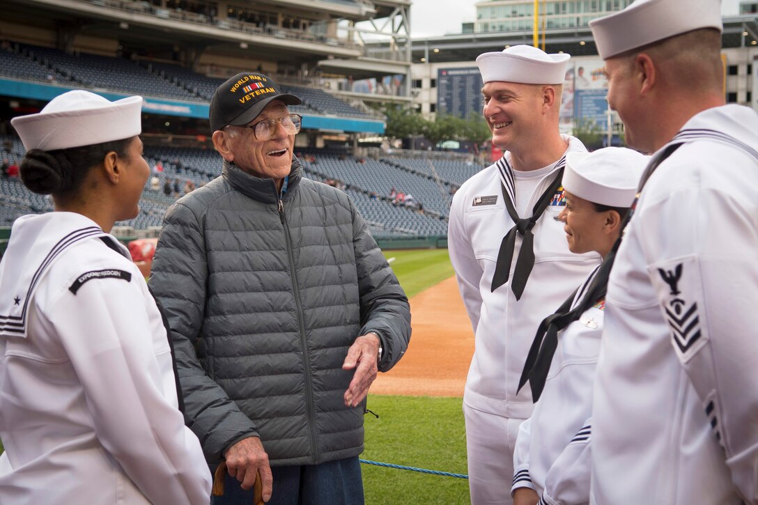 Four sailors and a veteran talk and laugh in a baseball field.