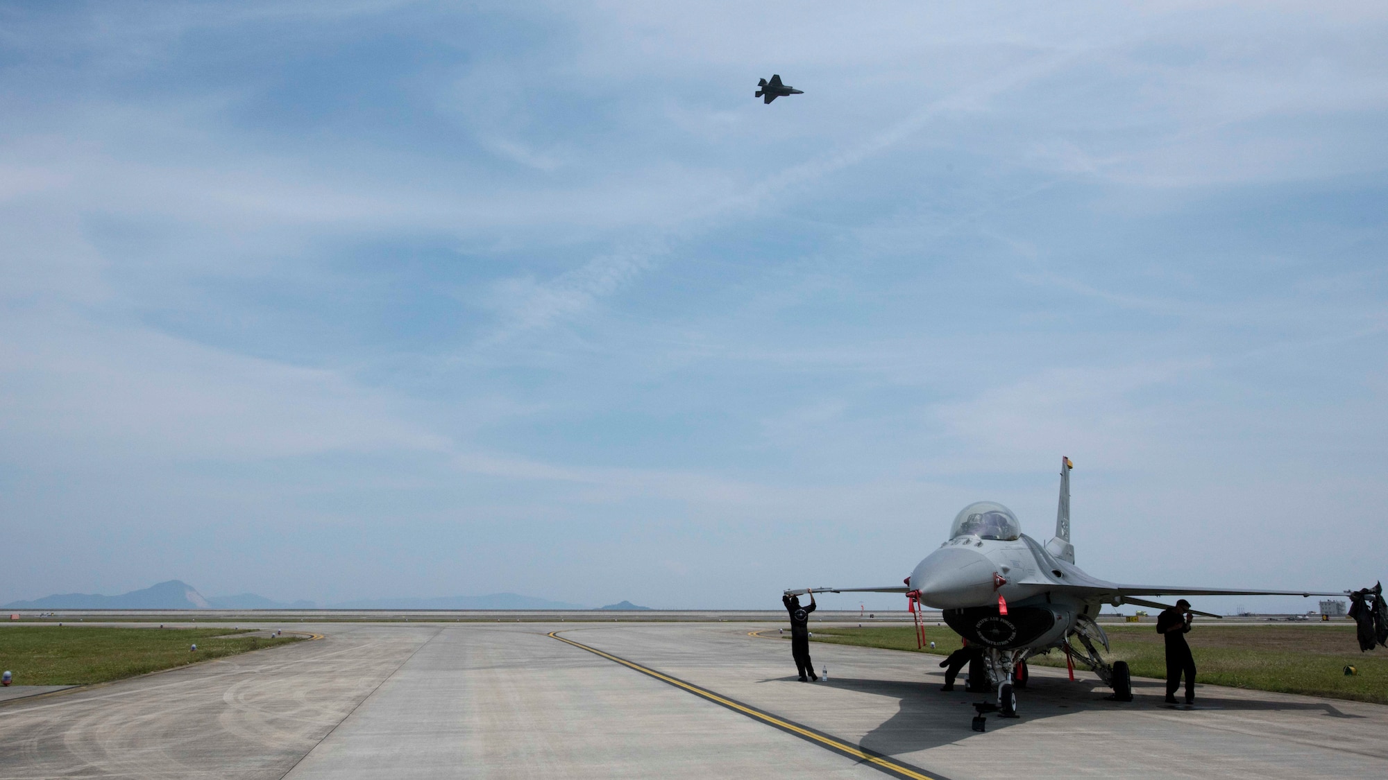 An F-35B Lighting II demonstration aircraft flies across the sky, as an F-16 Fighting Falcon sits on the runaway at the 43rd Japan Maritime Self-Defense Force – Marine Corps Air Station Iwakuni Friendship Day 2019 at MCAS Iwakuni, Japan, May 5, 2019. This event provides dozens of performances and attractions by American and Japanese members to showcase their aviation ability and talent. (U.S. Air Force photo by Senior Airman Collette Brooks)
