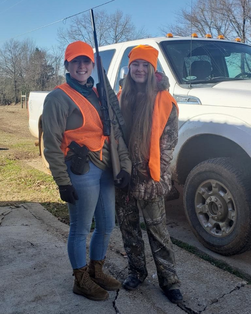 Two women in hunting clothes pose for a photo with a rifle.