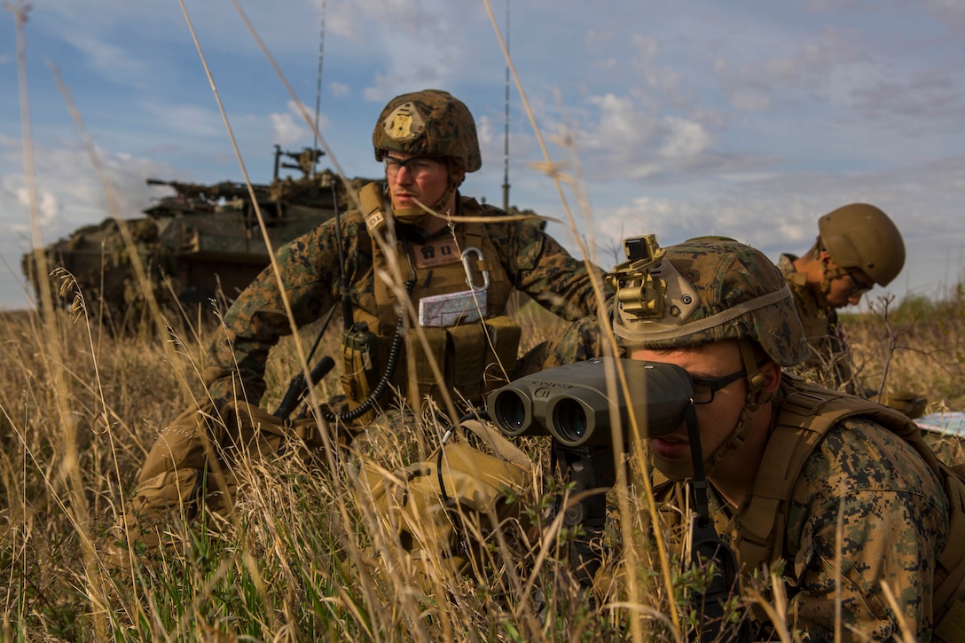 A service member looks through binoculars while lying in tall grass in front of an armored vehicle as others look on and check a map.