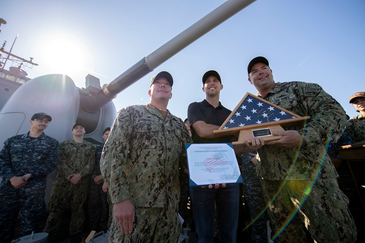 A civilian stands with sailors on the deck of a ship while holding a framed U.S. flag and a certificate. An MK 45 5-inch lightweight gun is in the background.