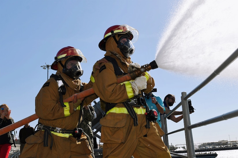 Two men in firefighter gear spray a fire hose off the deck of a ship as two men in the background take photos.