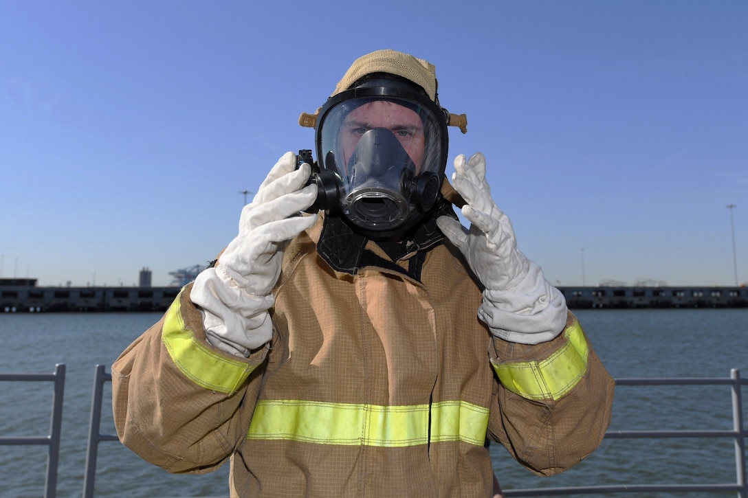 A man on the deck of a ship wearing a firefighting suit adjusts the mask he’s wearing with gloved hands.