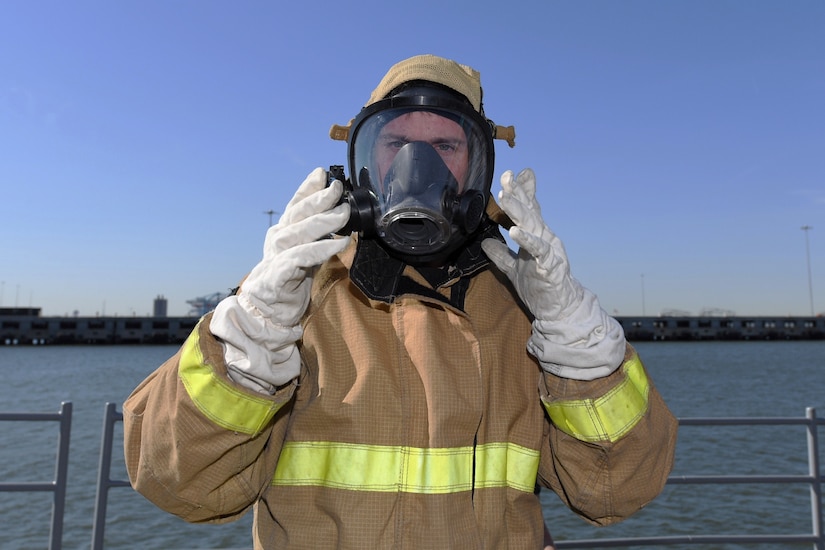 A man on the deck of a ship wearing a firefighting suit adjusts the mask he’s wearing with gloved hands.