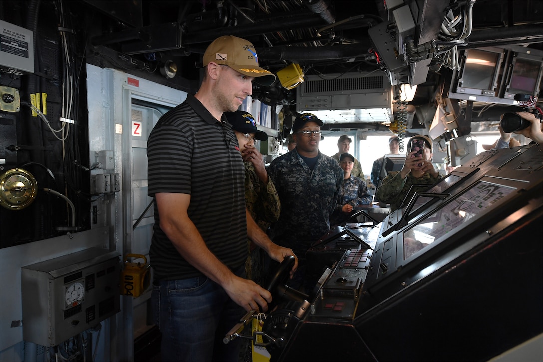 A civilian holds the wheel and looks at the controls on the bridge of a Navy cruiser as sailors look on.