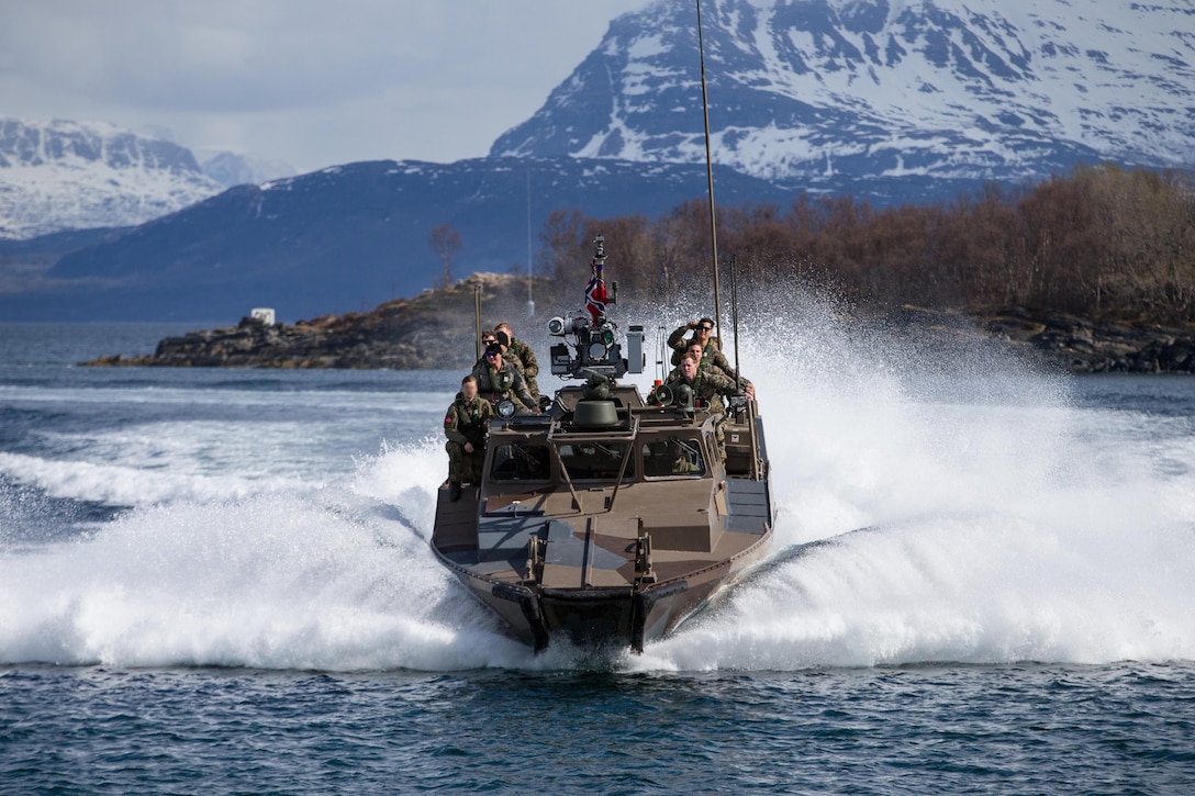 Marines speed across water on a boat with snow capped mountains in background.
