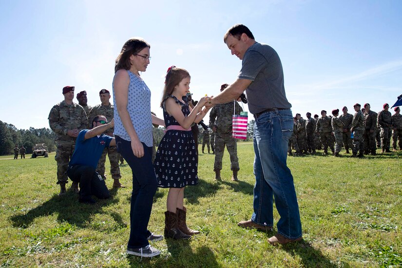 A young girl, standing beside her mom, puts a bracelet on a man in a field with soldiers around.