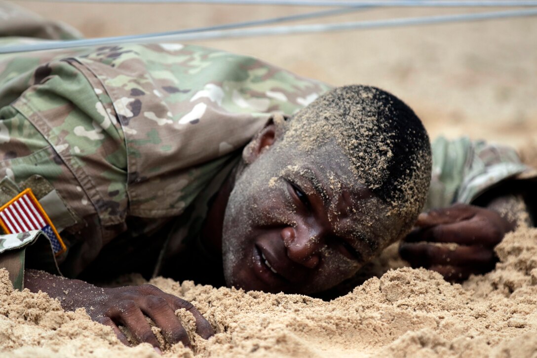 A soldier low-crawls in sand beneath an obstacle.