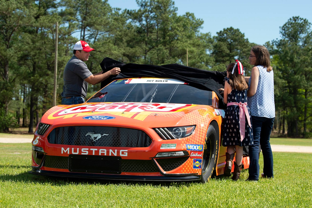 Three people remove the cover from an orange Ford Mustang race car.