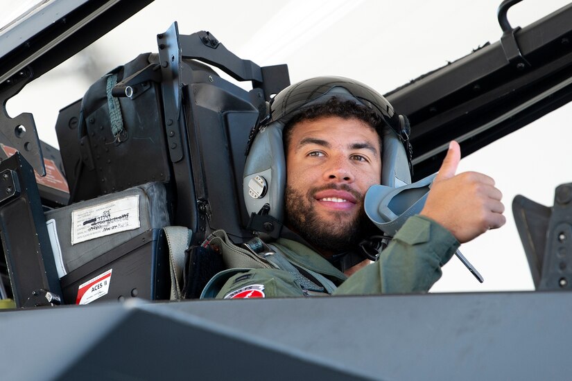 A man in flight gear and a helmet sits in a cockpit.