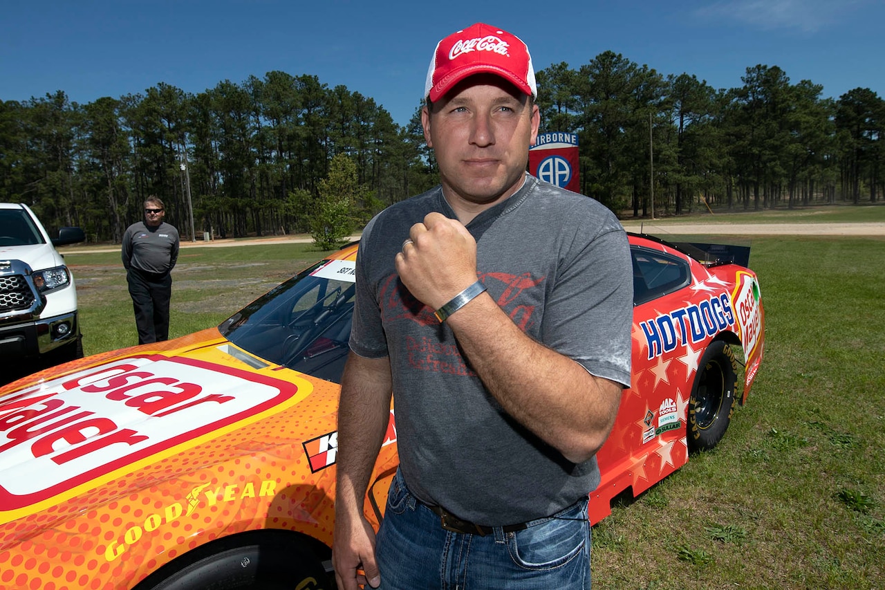 A man standing in front of a race car shows off a silver bracelet.