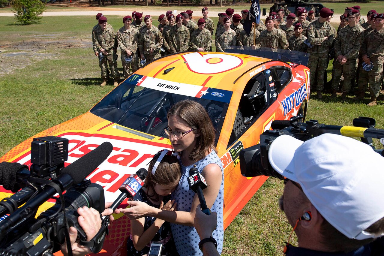 A mom and daughter standing in front of a race car talk to media as soldiers stand in the background.
