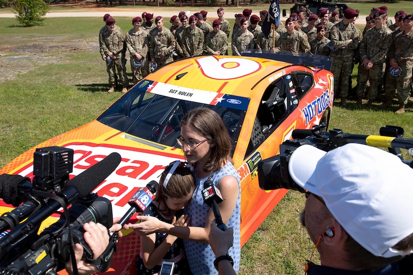 A mom and daughter standing in front of a race car talk to media as soldiers stand in the background.