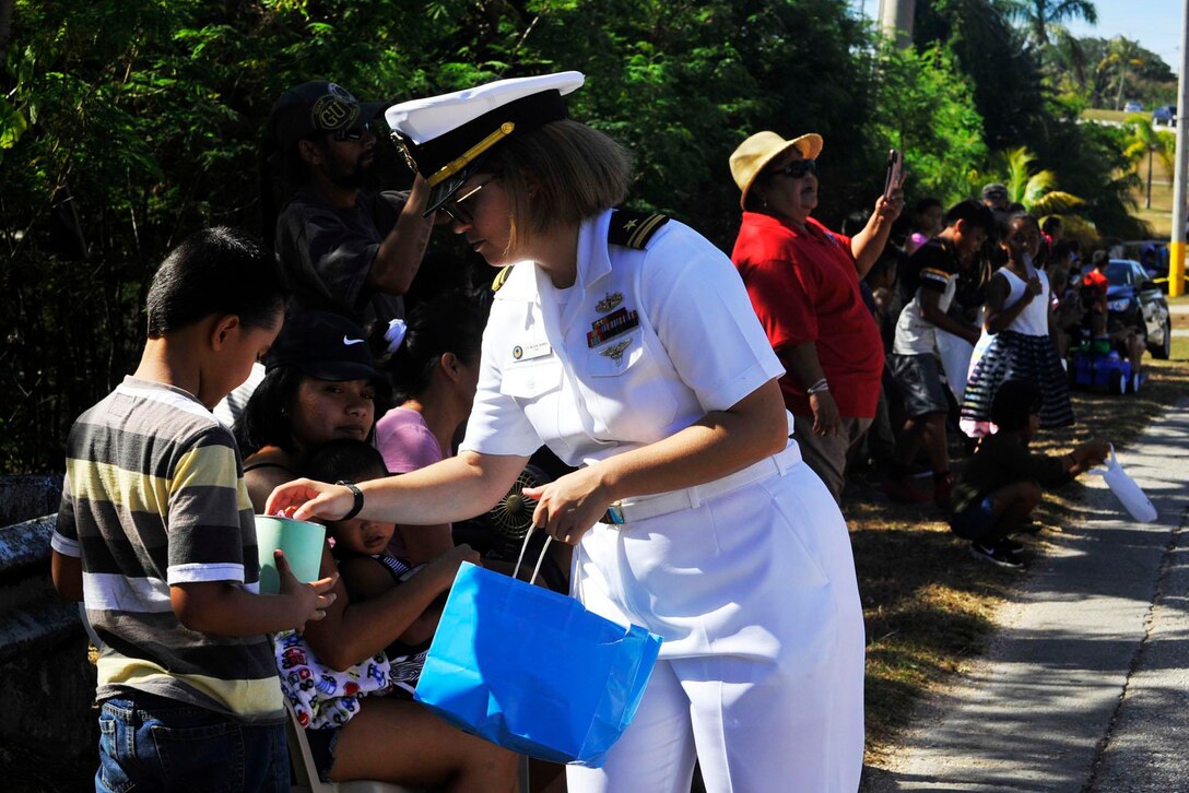 A sailor carrying a paper shopping bag places candy in a container a child is holding as others line a sidewalk to watch a parade.