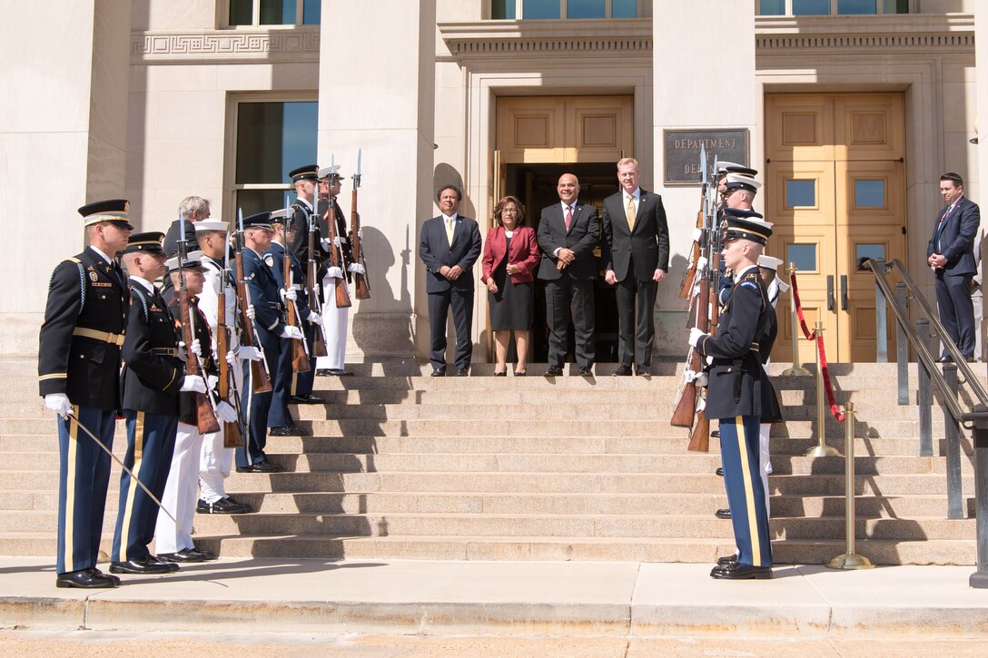 Four defense leaders stand at the top of some steps while a group of service  members stand  in a line on both sides of them.