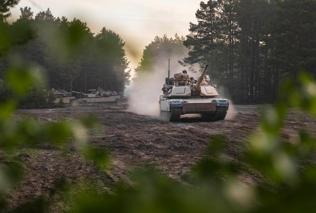 A tank with several soldiers aboard kicks up dust as it rolls down a path lined with trees.