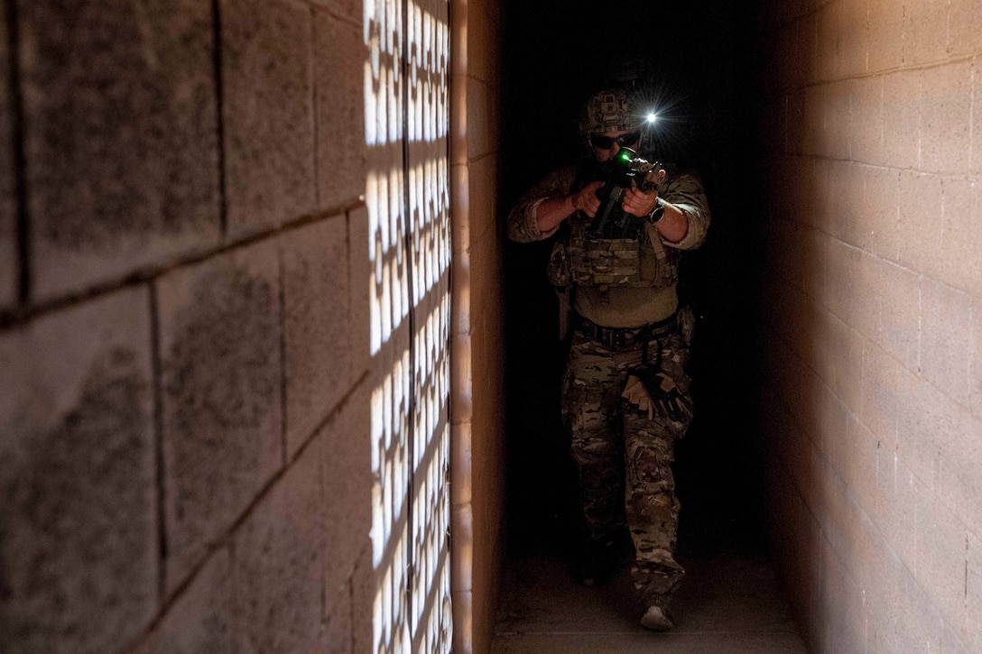 A sailor wearing a helmet with an attached light emerges from a dark hallway aiming a rifle.