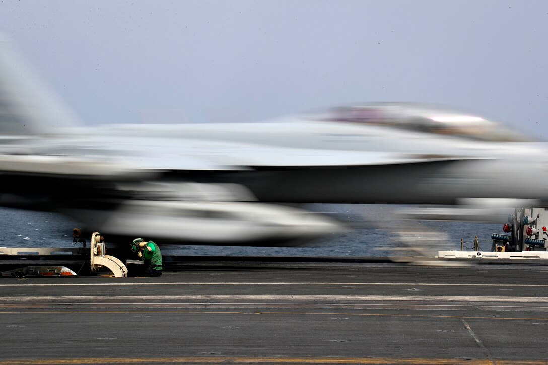 A fighter jet races past a sailor who is sitting and working on the flight deck of an aircraft carrier.