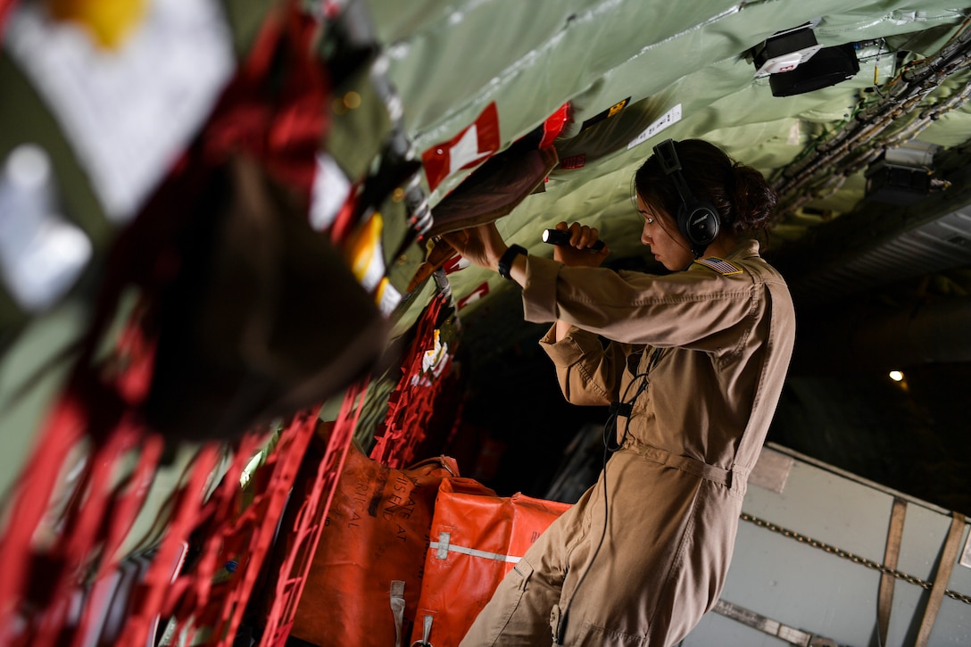 An airman uses points a flashlight at a panel in the wall of an aircraft.