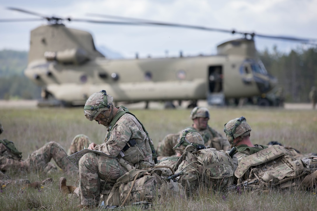 A group of soldiers in camouflage uniforms and helmets sits in the grass as a helicopter awaits in the distance.