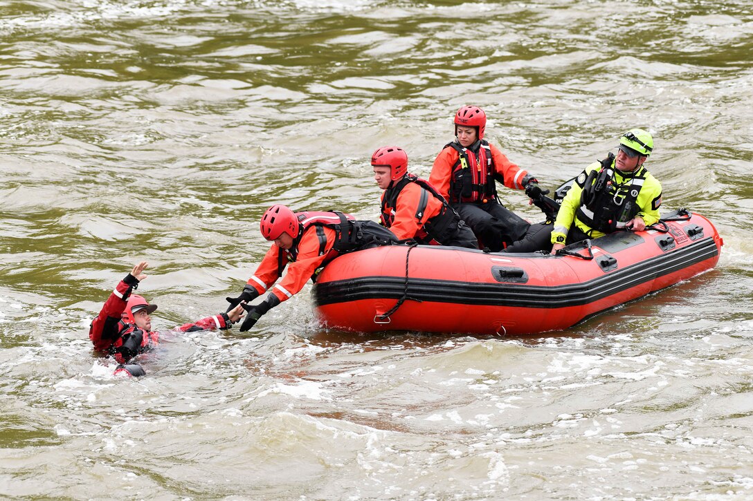 Airmen wearing vests, waders and helmets form a chain inside a raft to reach and rescue another airman in a river.