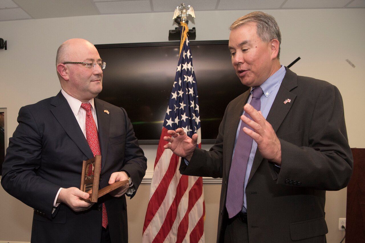 A man speaks and uses hand gestures as another man listens.