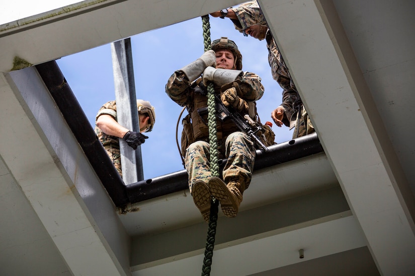 A Marine sits at the opening of a square hole holding a rope between his arms and legs.