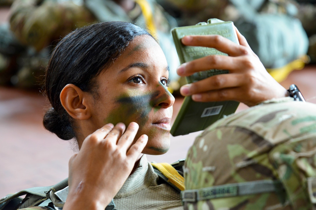 A soldier applies pant onto her face will looking at a compact mirror.