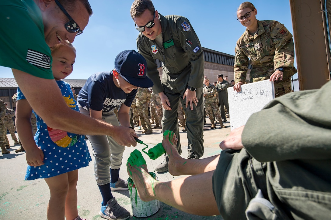 An adult and a child paint a pilot’s feet green with a roller as airmen and another child look on.