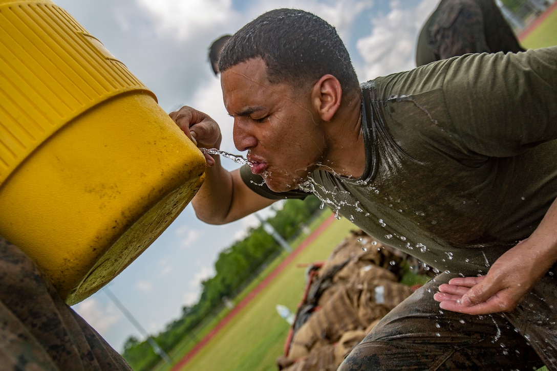 A Marine kneels to dispense and drink water from a large yellow jug held by another Marine.