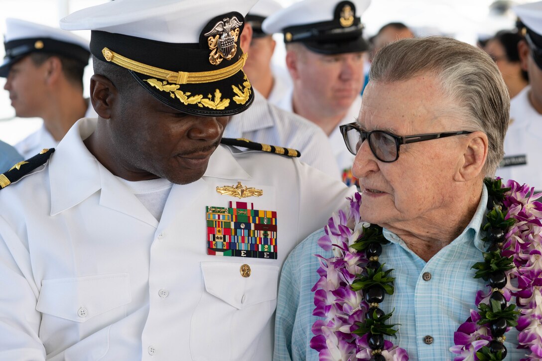 A sailor speaks with a veteran as several other sailors in dress whites stand in the background.