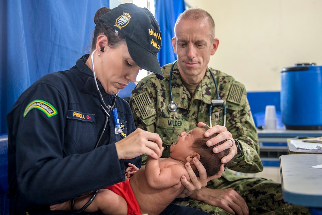 A Brazilian military doctor holds an infant as a Navy doctor puts his hand on the baby's head.