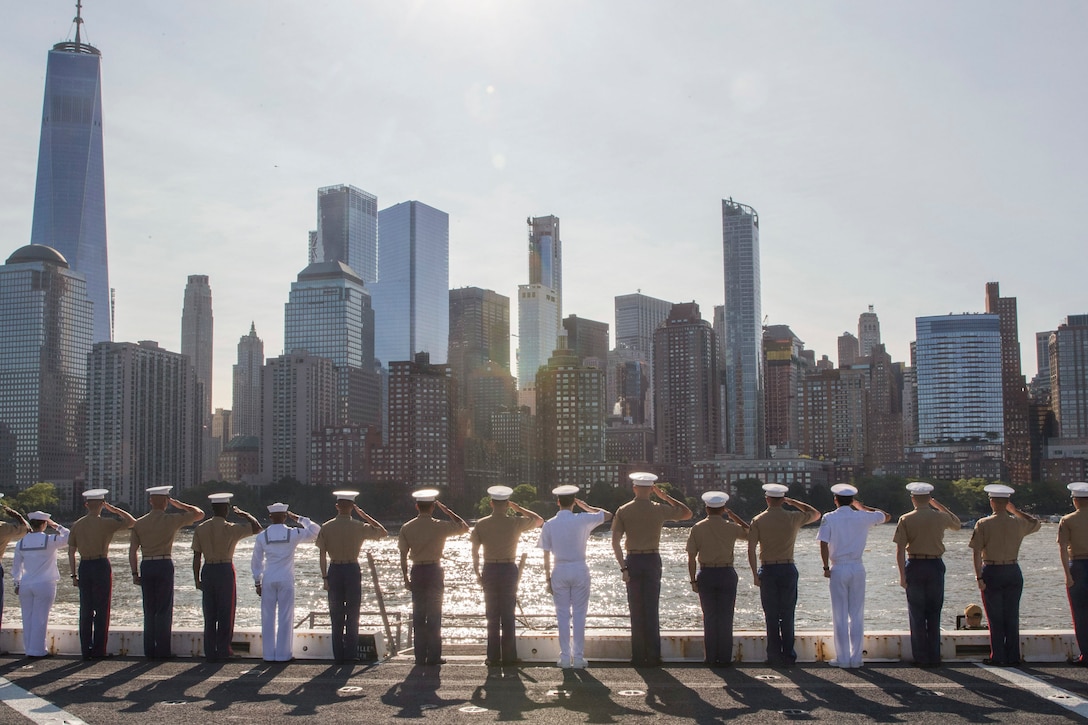 Sailors and Marines salute from a ship's deck while facing the New York City skyline.