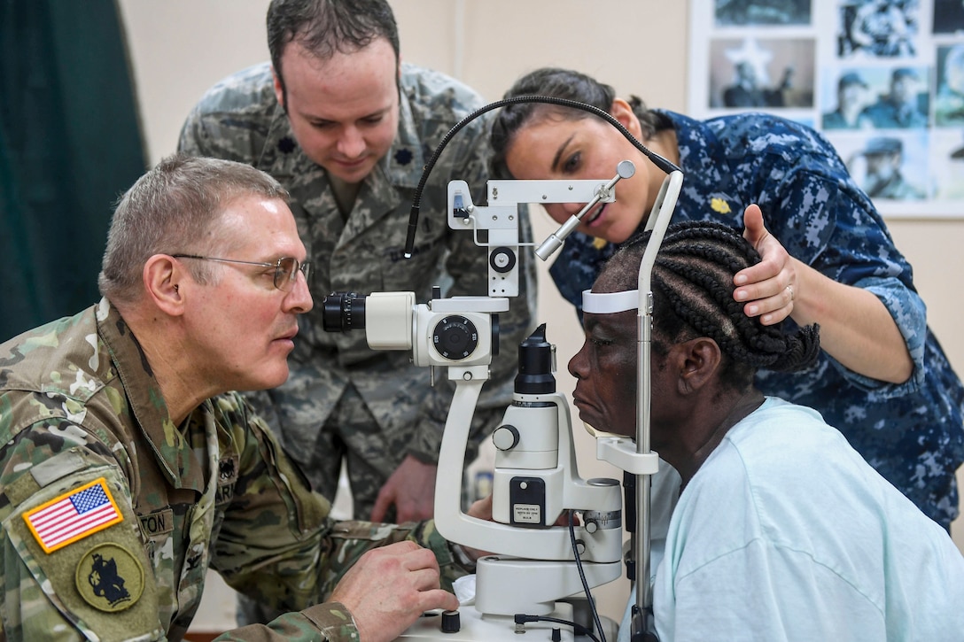 Three service members examine a patient whose head is resting on an ophthalmology device.