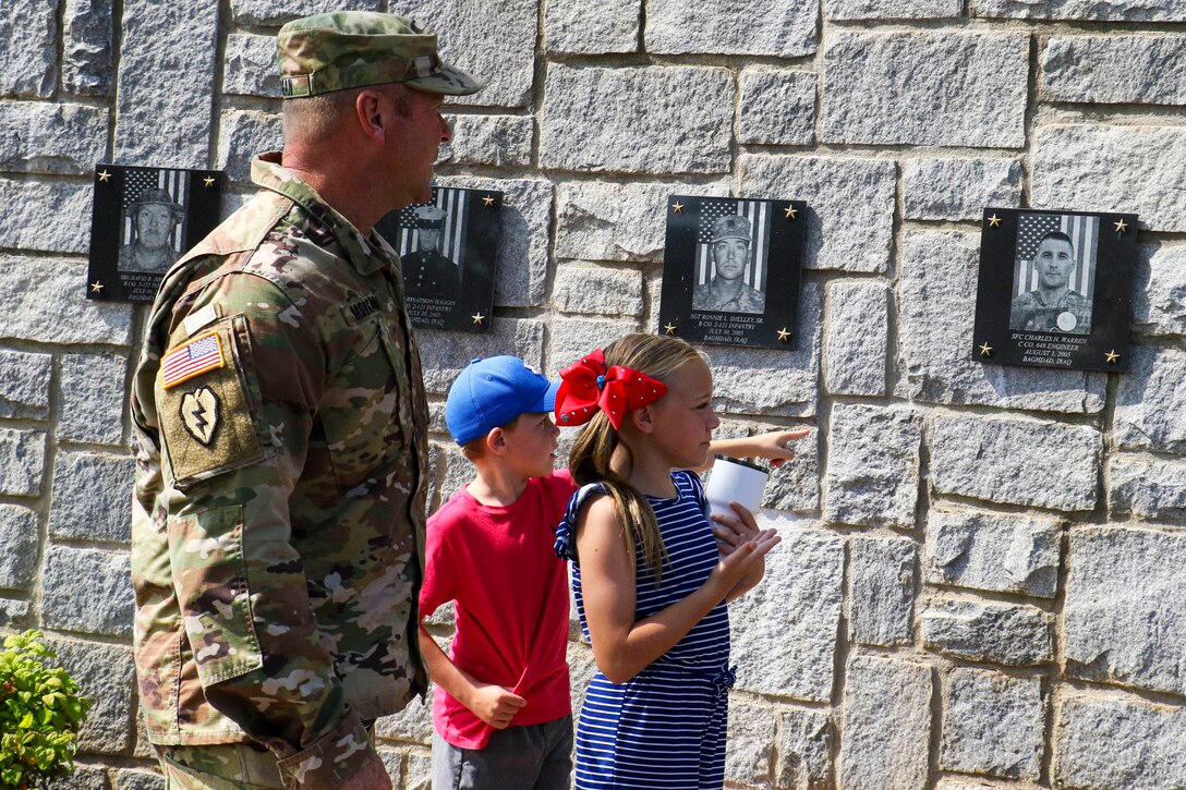 A soldier and two children point to pictures of fallen soldiers on a wall.