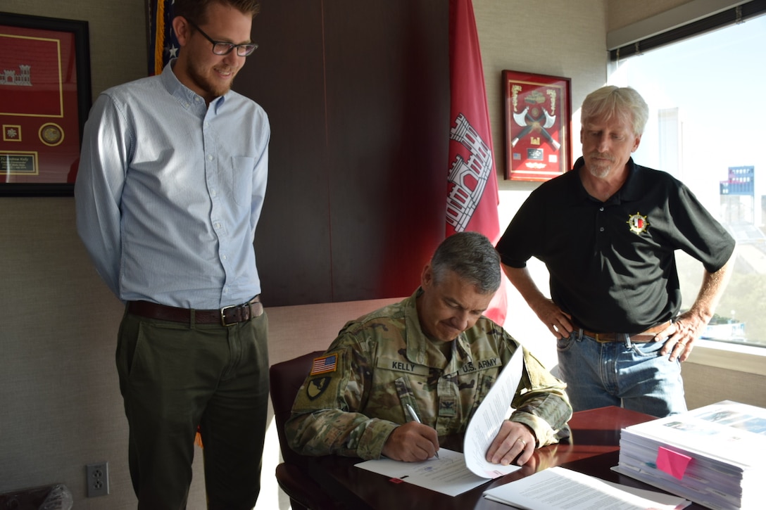 Col Kelly sitting at his desk signing a document while two Jacksonville district employees stand by his sides