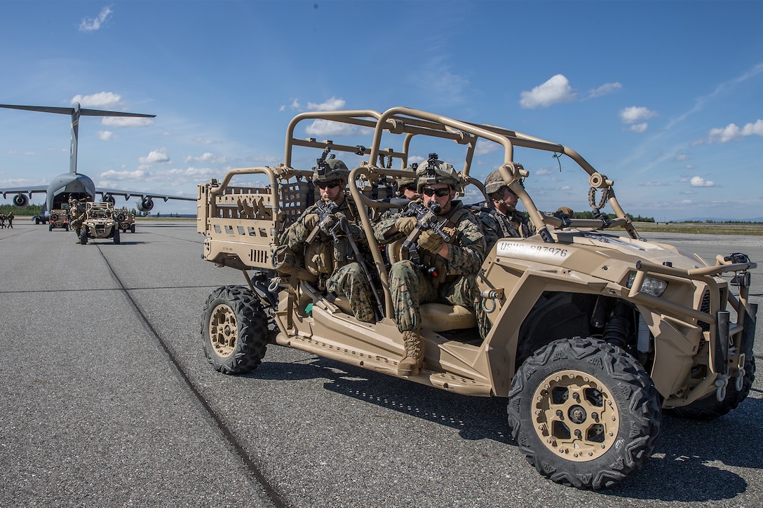 Marines ride in an all-terrain vehicle with an aircraft in the background.