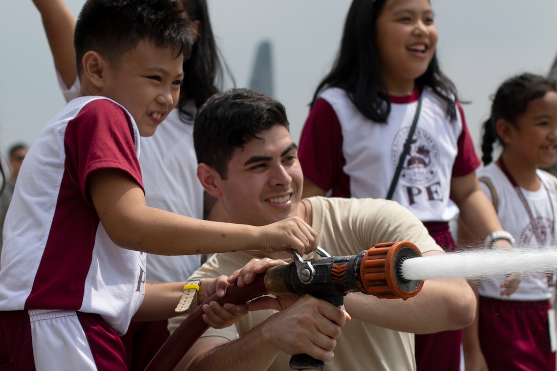 A smiling airman helps a smiling child hold a spraying fire hose as other kids watch.