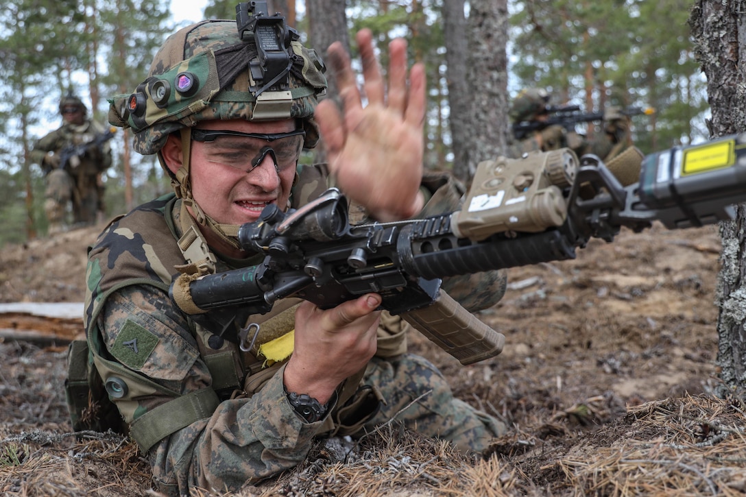 A Marine wearing a helmet and goggles checks his rifle while on the ground in a forest in firing position.