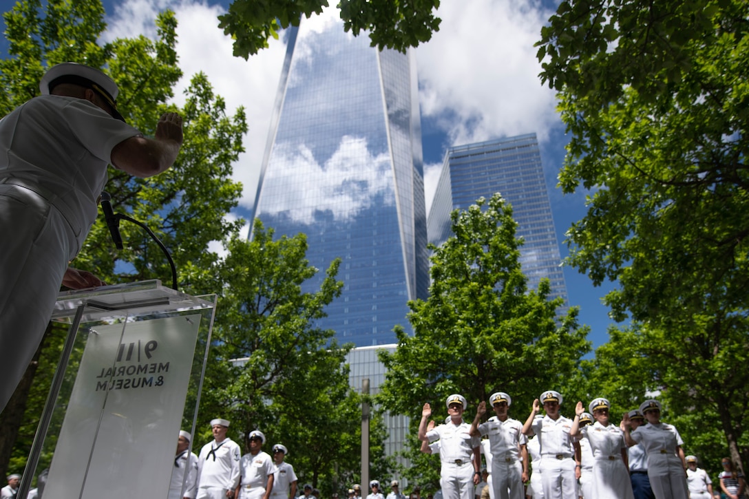 A naval officer stands behind a podium raising his right hand as several sailors in front of him raise their hands near New York’s Freedom Tower.