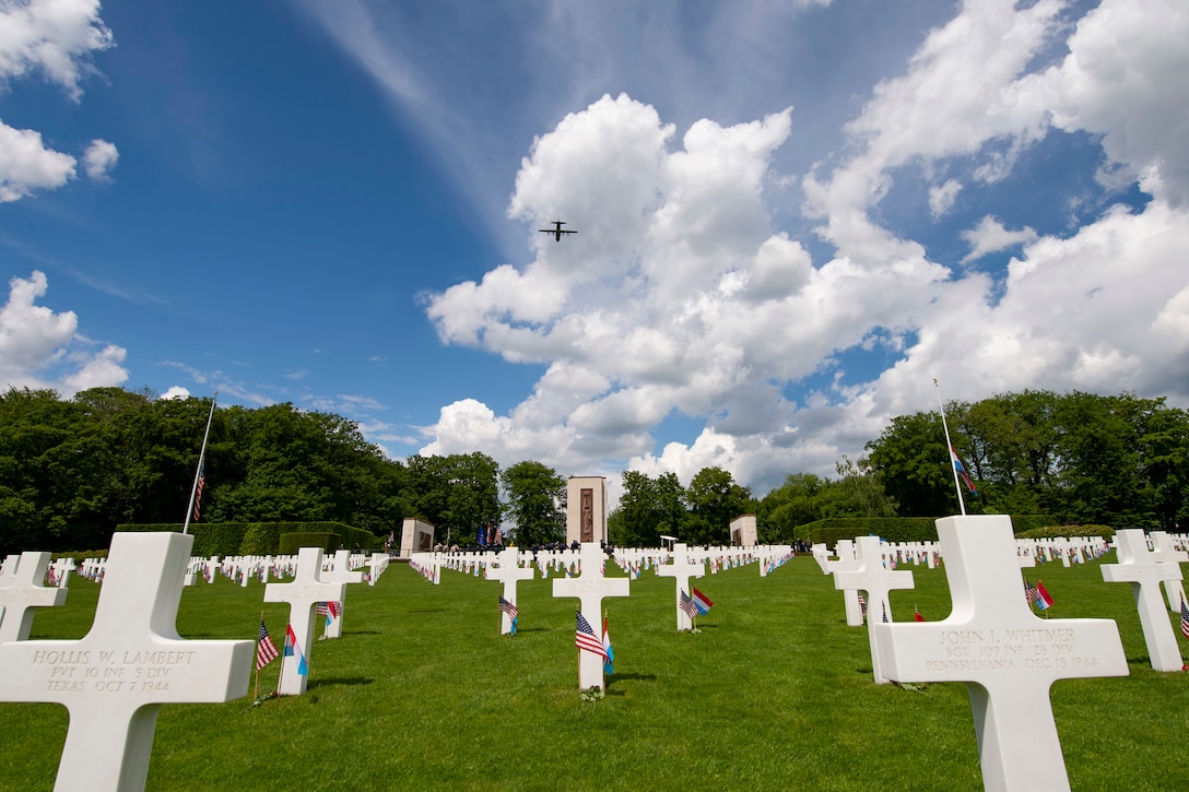 An aircraft flies above headstones amid clouds and blue sky.