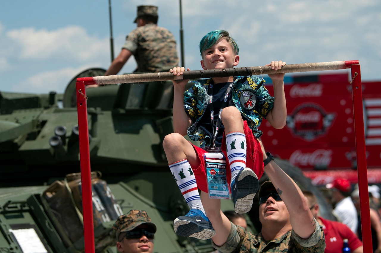 A boy does a pullup with the help of a service member.