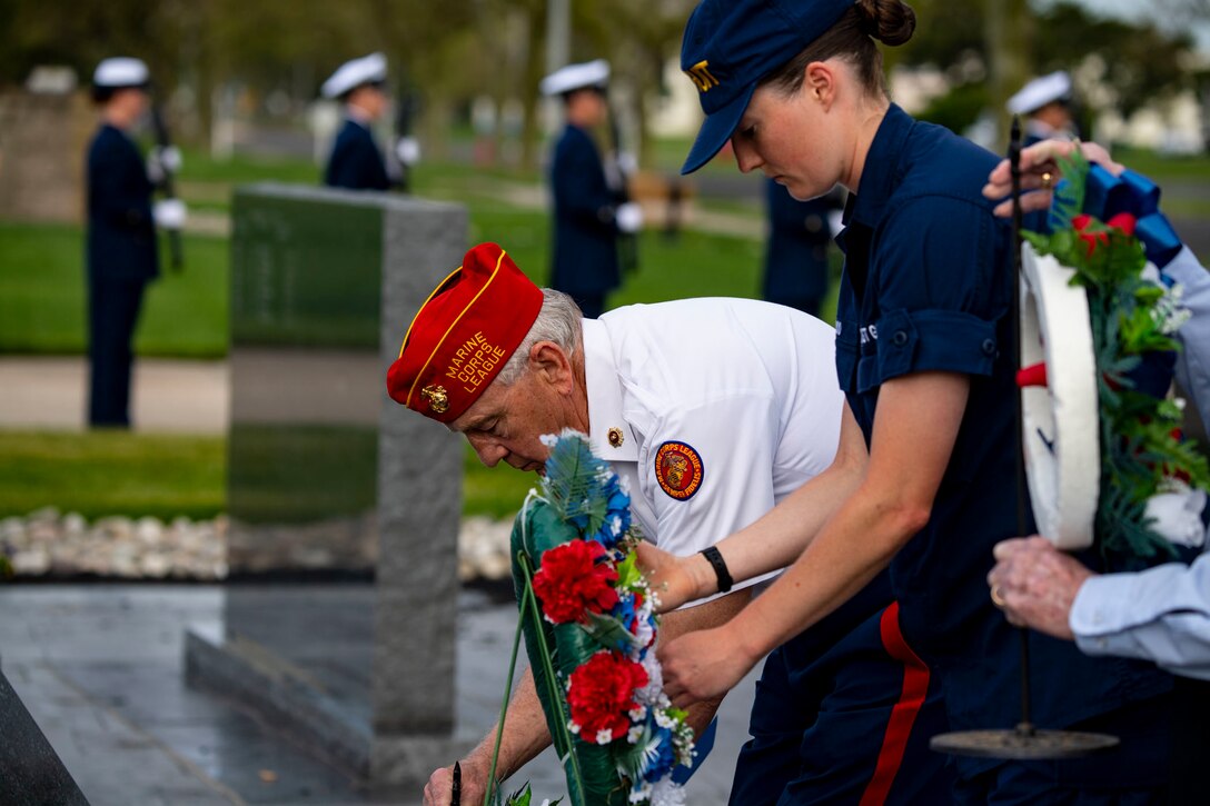 A coast guardsmen holds a wreath while a veteran writes a note at a memorial.