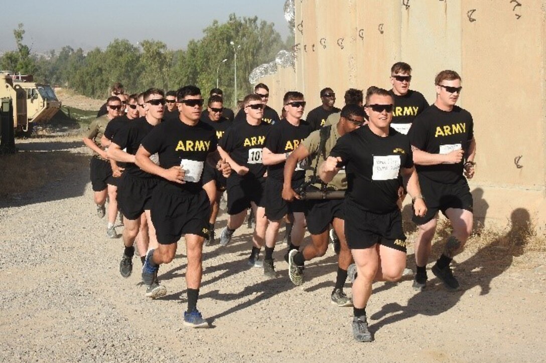 A group of soldiers run past a wall and some vehicles.