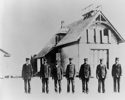 A black and white photo of a crew of black surfmen. They are wearing suits and standing in front of the lifesaving station.