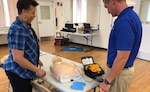 Woman and man stand at a table with a CPR training body and AED.